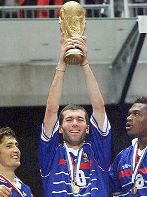 Soccer player in blue jersey holding FIFA World Cup trophy above head with teammates beside him