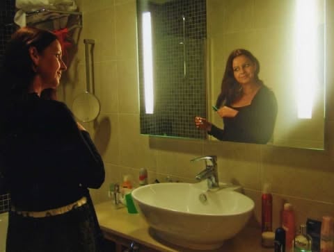Person with red hair standing at a bathroom sink, reflected in an illuminated mirror with green tile backdrop