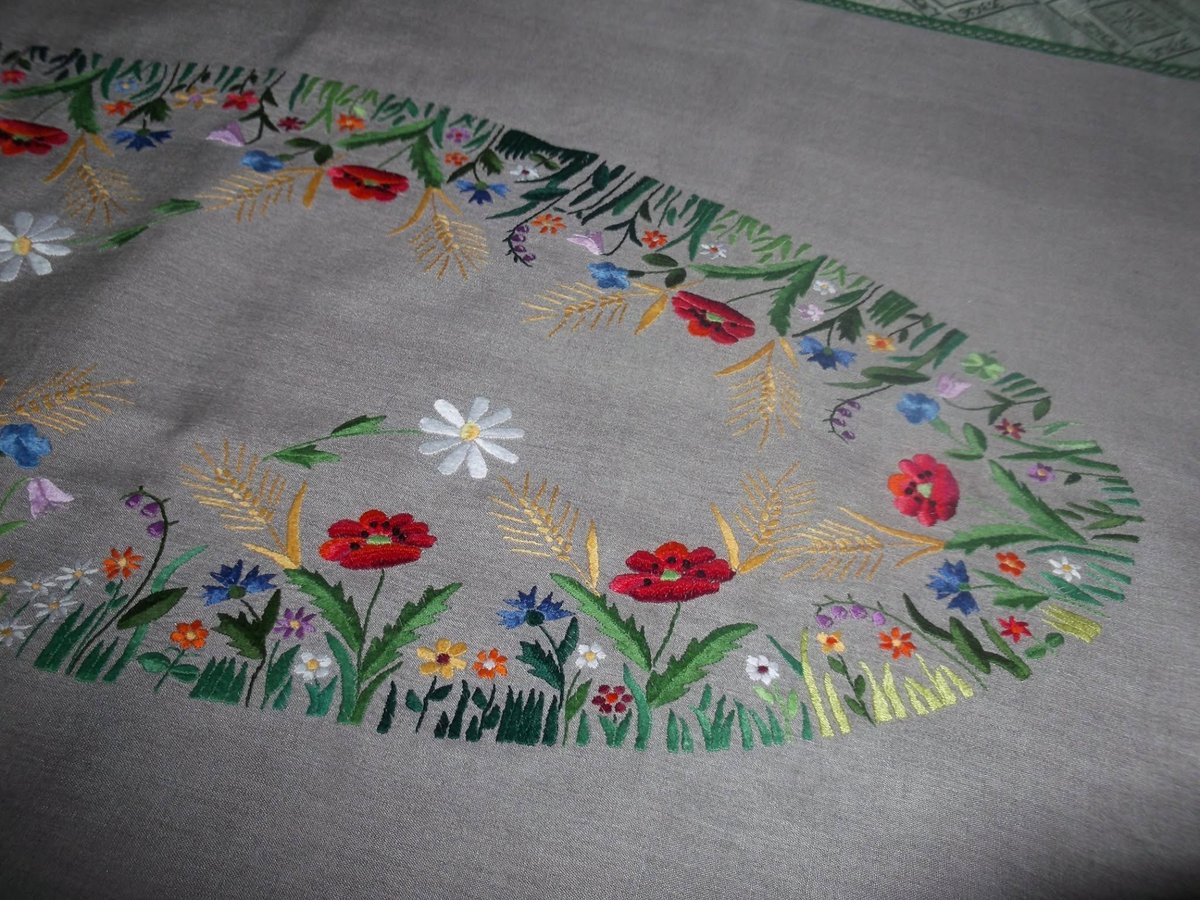 Embroidered fabric with colorful floral and wheat designs along the scalloped edge on gray linen