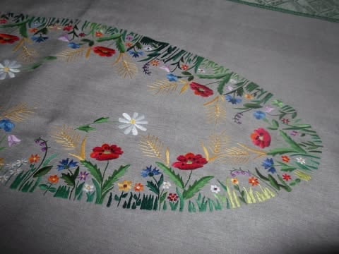 Oval embroidered table runner or placemat featuring colorful flowers including red, blue, and white blooms with green leaves and wheat stalks on beige linen fabric