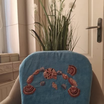 Blue chair with reddish-brown embroidered decorative pattern, tall dried grass plant in blue ceramic pot on back