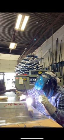 Worker in blue protective gear welding metal parts in an industrial warehouse with bright overhead lighting