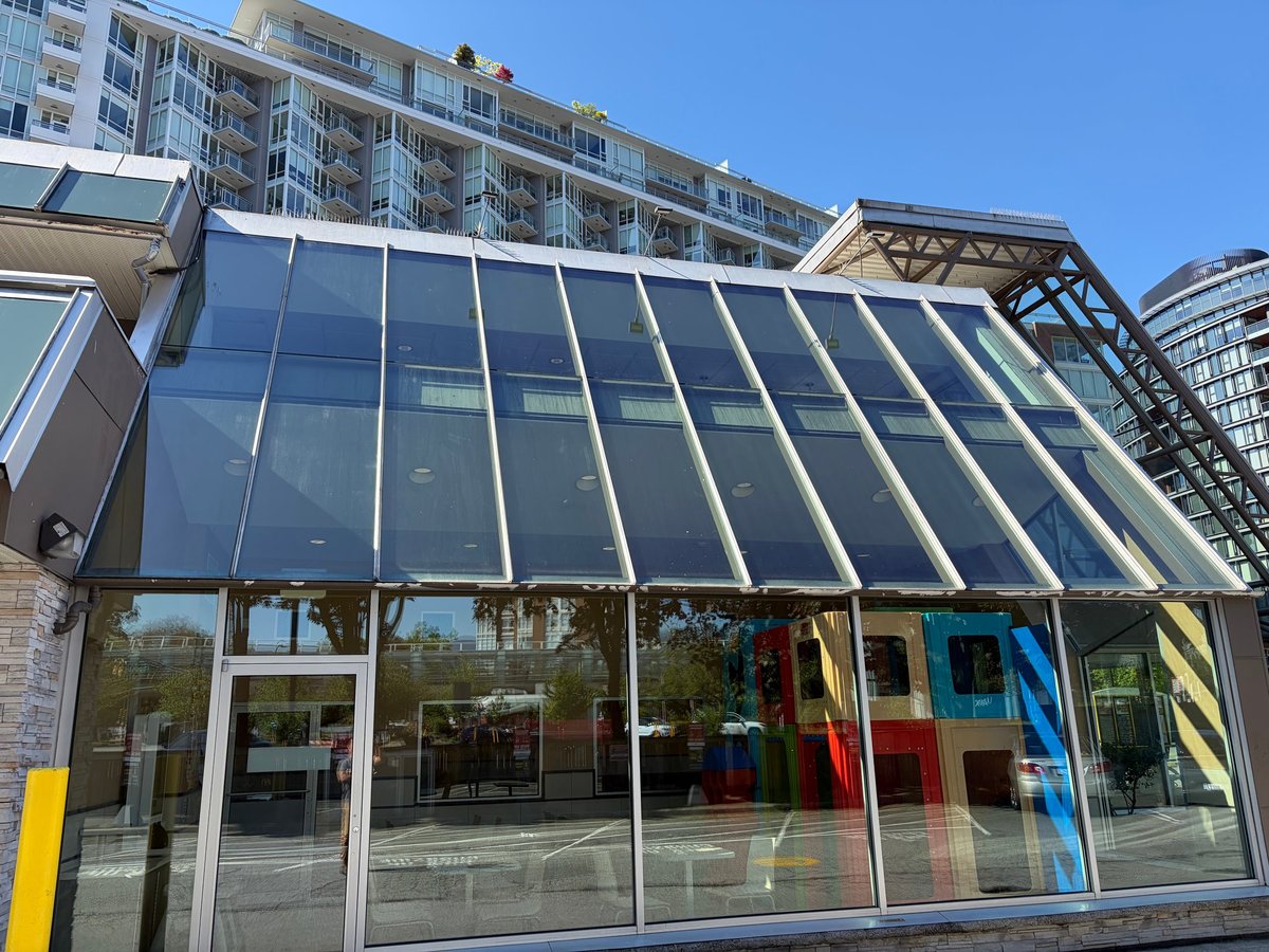 Modern glass storefront with sloped metal roof beneath residential buildings under clear blue sky