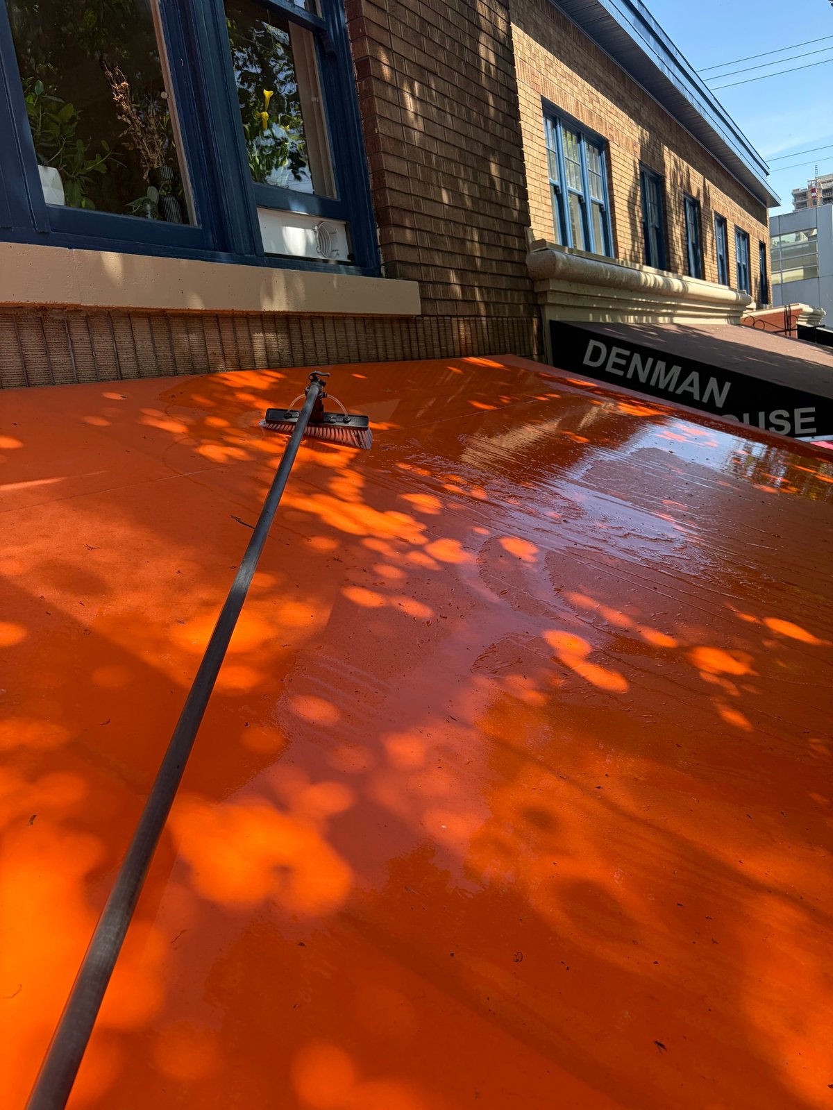 Bright orange-red awning covering a storefront with blue windows and brick building, casting leaf shadows