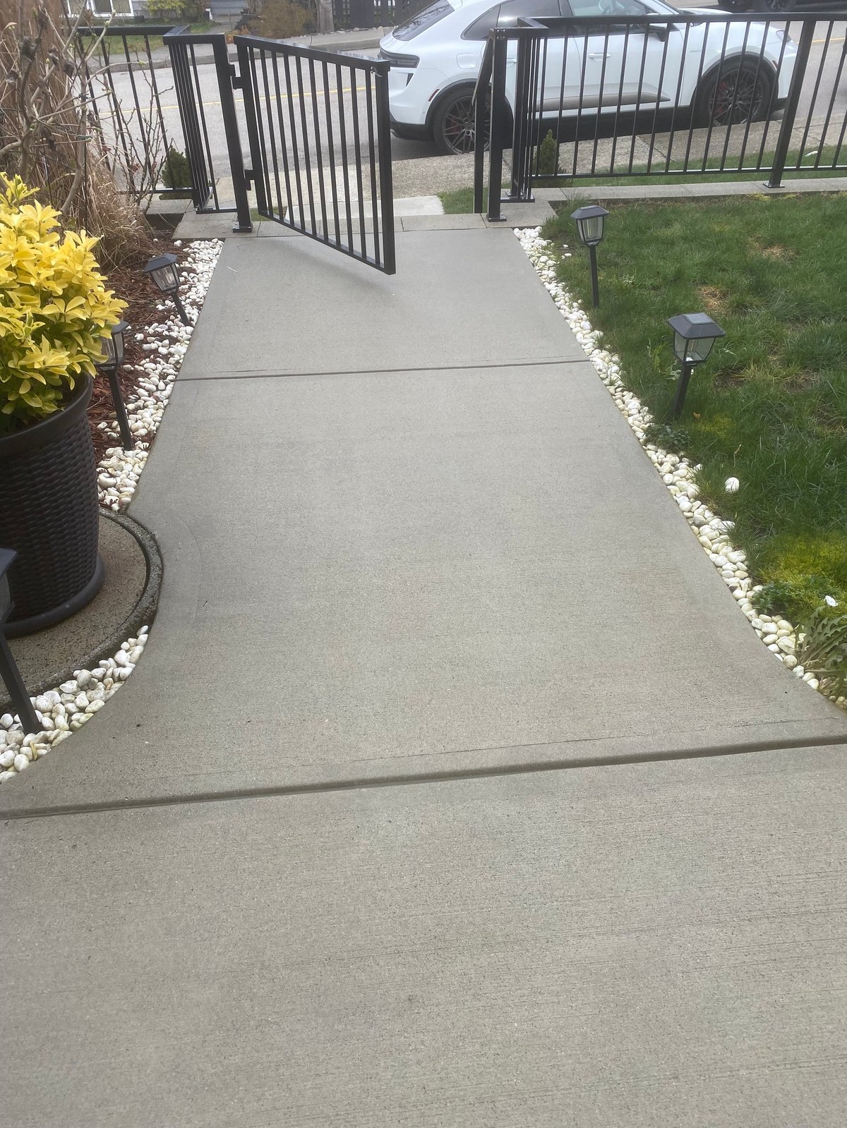 Concrete driveway and walkway leading to open black metal gates with white vehicle and landscaping visible