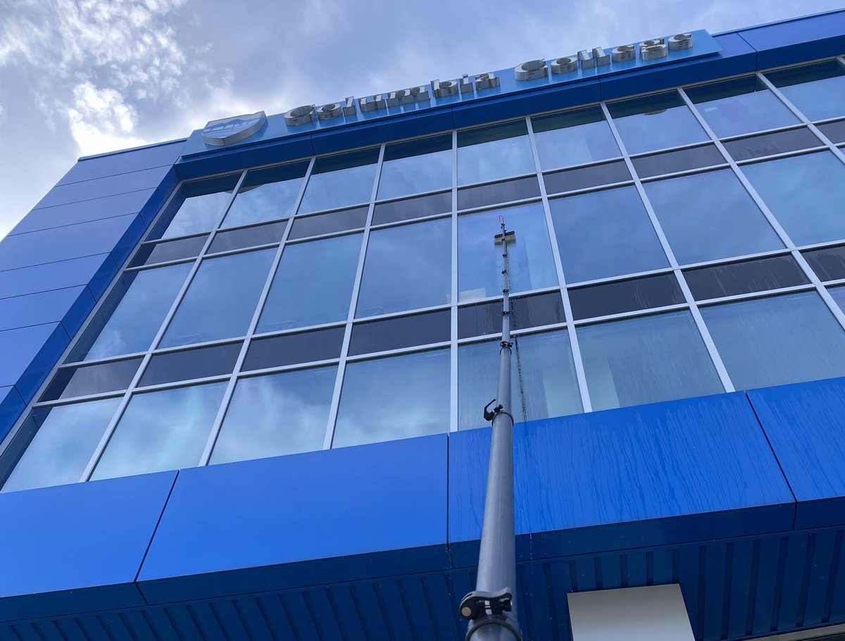 Modern blue and glass building exterior with large gridded windows and metal antenna pole against cloudy sky