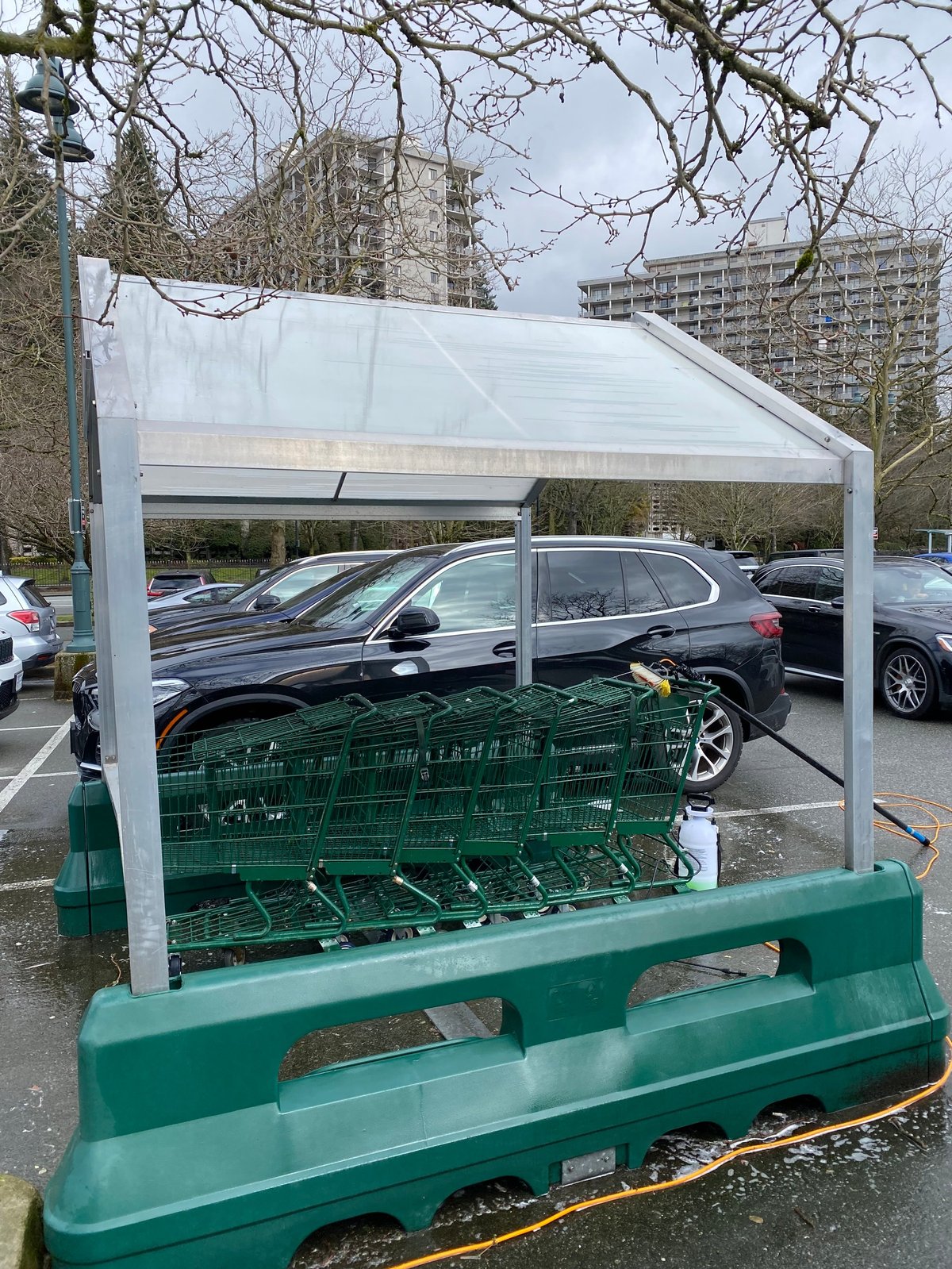 White canopy covering stacked green shopping carts in a parking lot with cars and apartment buildings in background