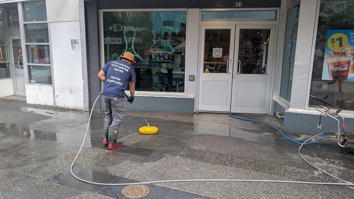Worker in blue shirt pressure washing storefront pavement with yellow equipment and white hose.