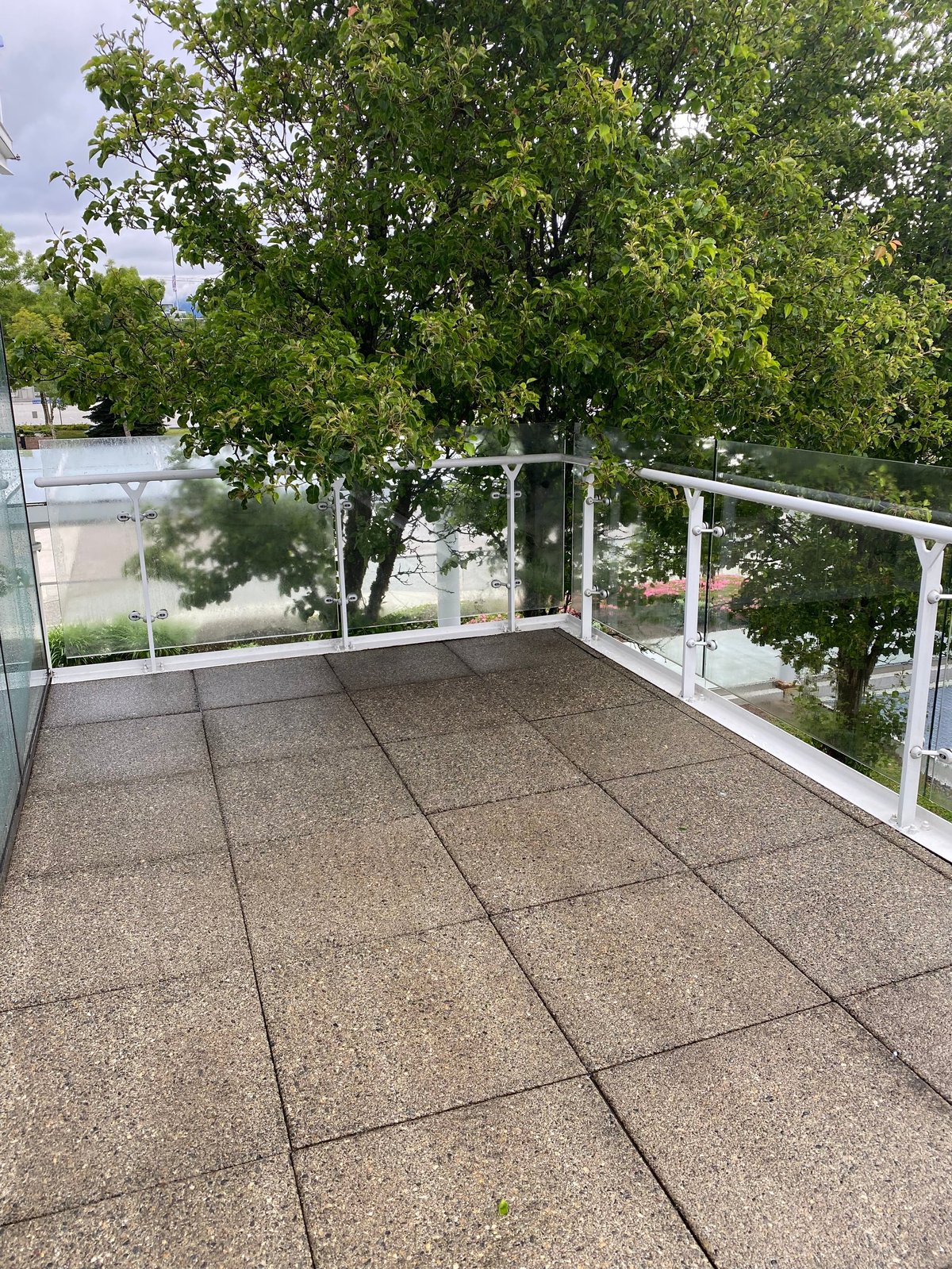 Modern balcony with gray paved flooring, glass and white metal railing, overlooking trees and water landscape