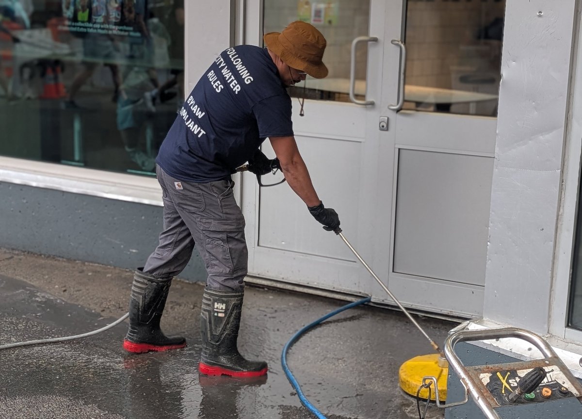 Worker in dark uniform pressure washing storefront with yellow equipment and blue hose on wet pavement