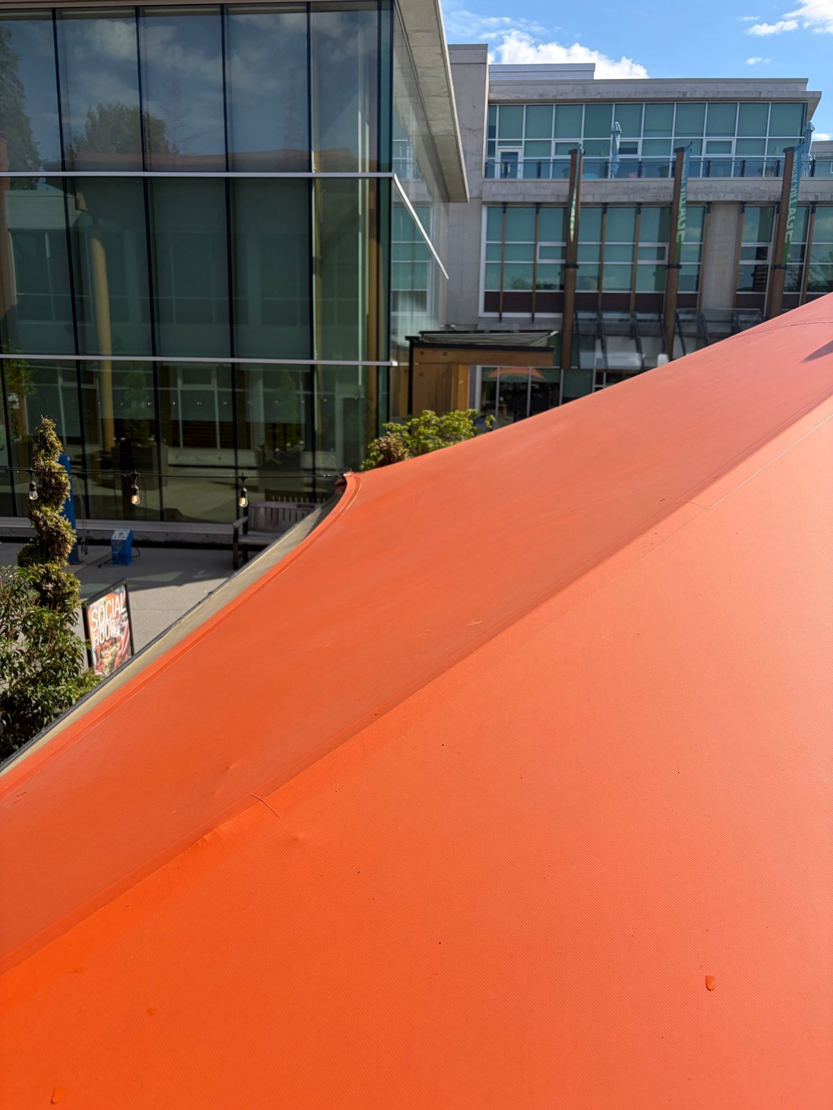 Large orange curved metal structure with modern glass buildings and courtyard in background under blue sky