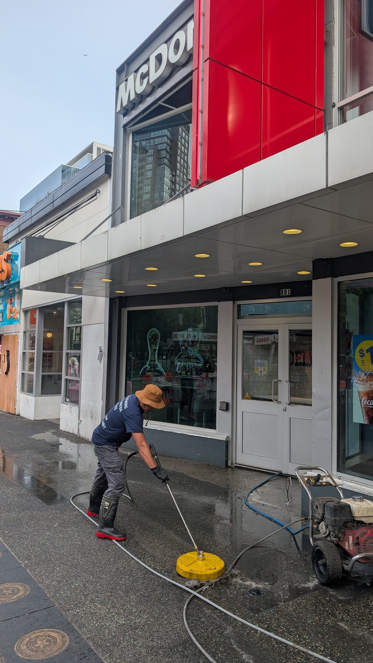 Worker pressure washing storefront of McDoors building with yellow nozzle on wet pavement
