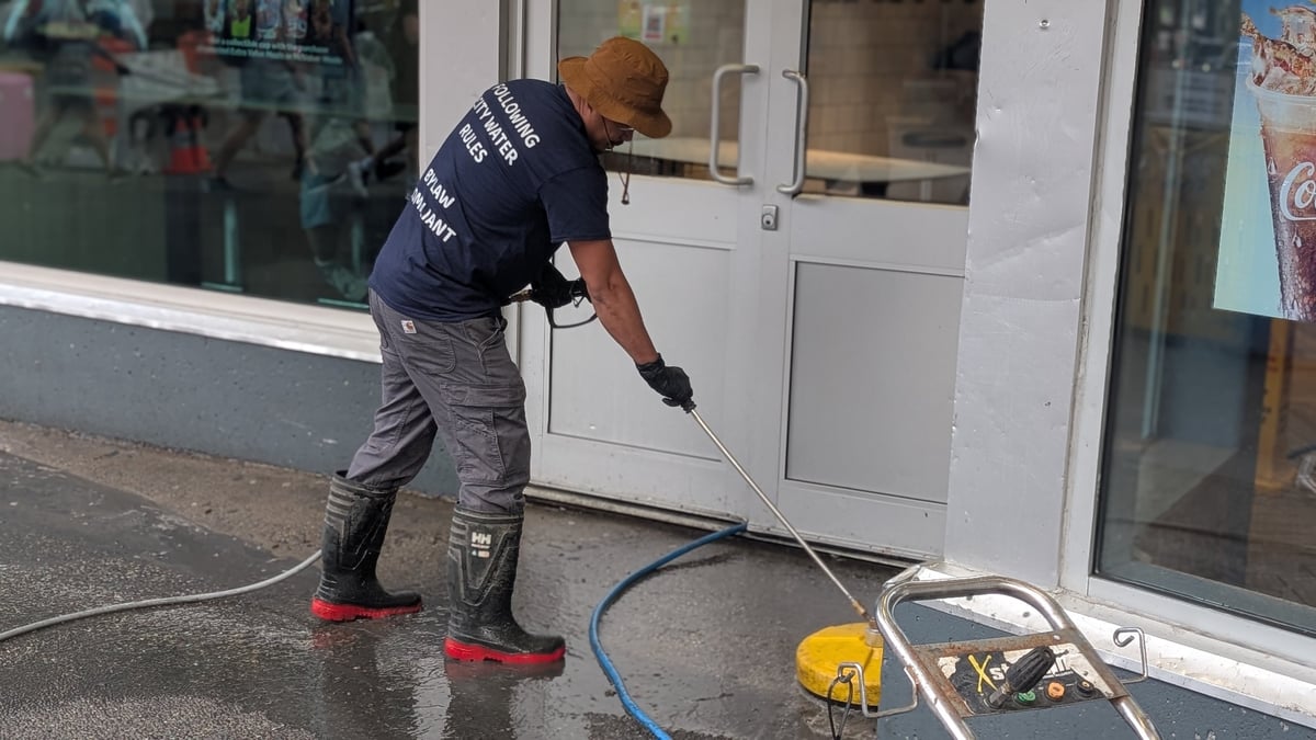 A person in dark work uniform sprays water on a storefront door and window using a pressure washer hose.