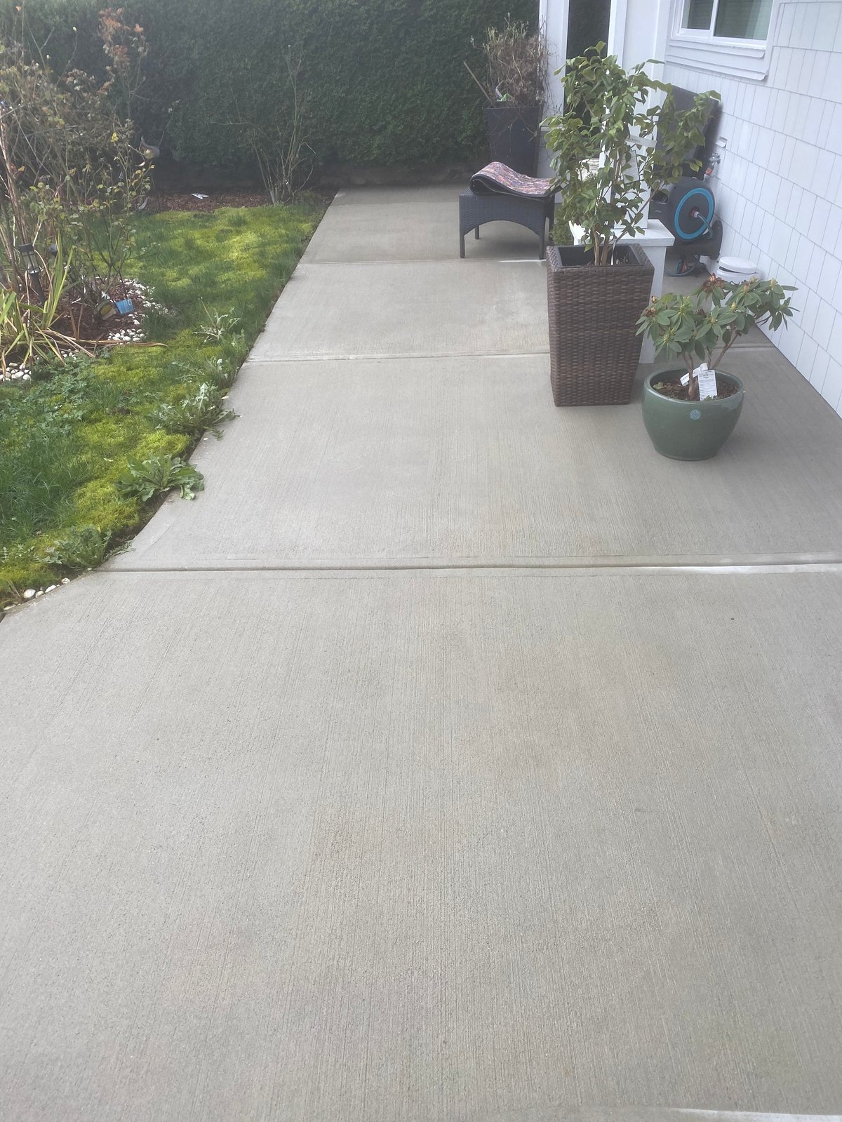 Concrete patio along white house with potted plants, lawn and hedges visible on left side