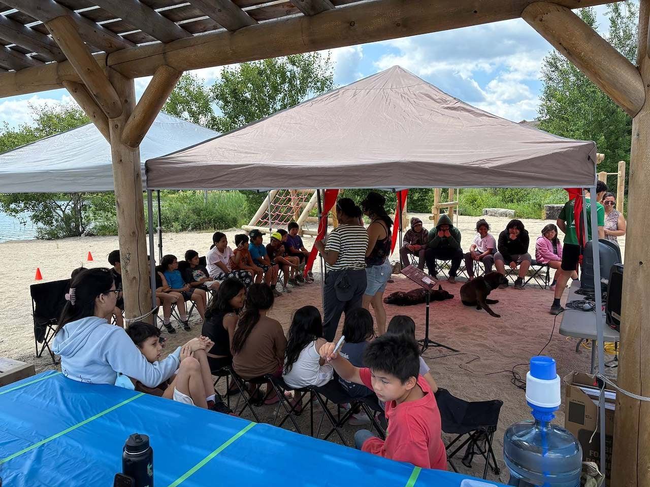 Group of people gathered under a large tent canopy at an outdoor event near a beach, with water and trees visible in background