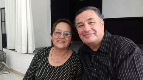 Two people smiling at camera indoors. Woman on left wears polka dot top and glasses; man on right wears dark pinstriped shirt