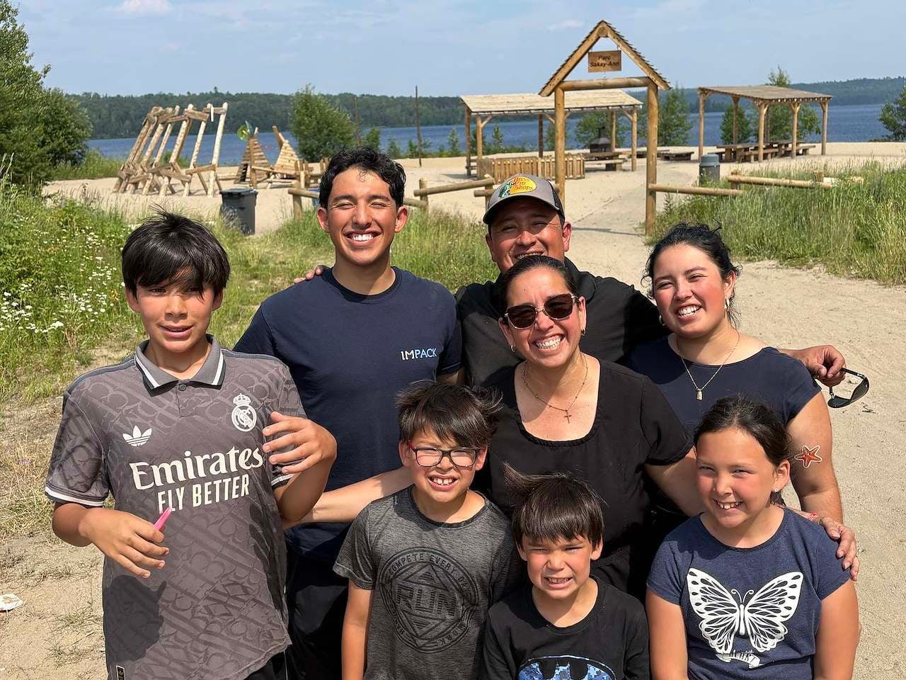 Group of six smiling people posing together on a sandy beach with playground structures and water visible in the background