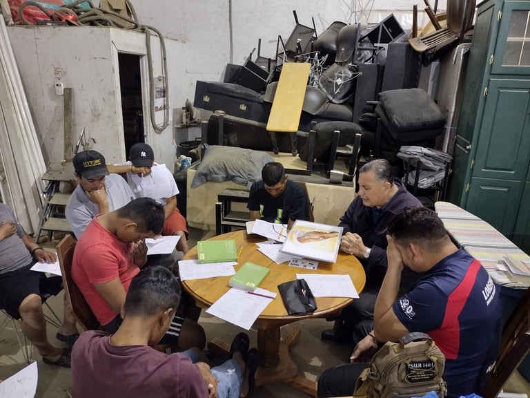 Group of people gathered around a wooden table in a cluttered workshop, reviewing documents and materials together