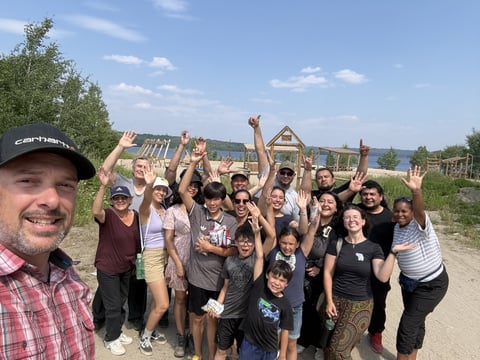 Large group of people cheering with raised arms outdoors on a rural dirt road with farm buildings in the background