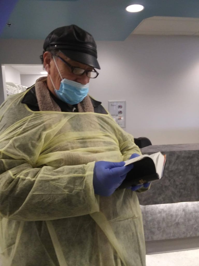 Person wearing protective gear including yellow gown, blue gloves, black cap, and face mask, holding a clipboard indoors