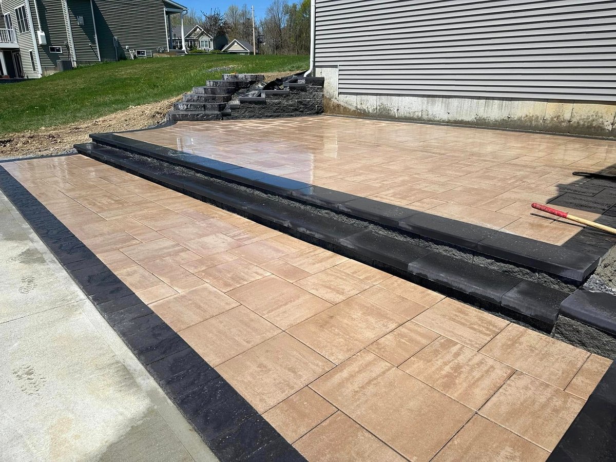 New paver patio under construction with black border edging and tan-colored pavers, residential backyard with siding home visible