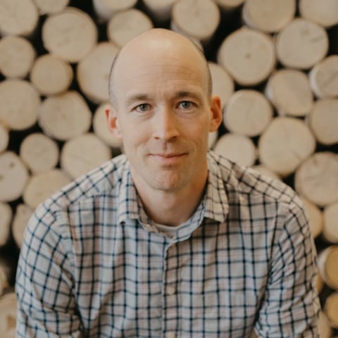 A bald man in a blue and white plaid shirt smiling at the camera with stacked logs in the background