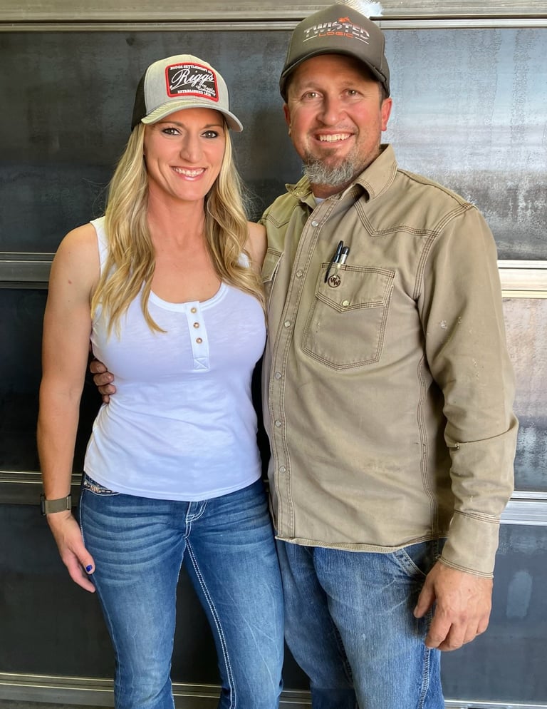 A man and woman standing together wearing casual clothing and branded baseball caps, smiling at the camera in front of a metal surface.