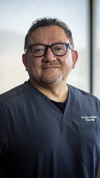 Professional headshot of a man with glasses wearing dark medical scrubs with embroidered text, against a light blurred background.