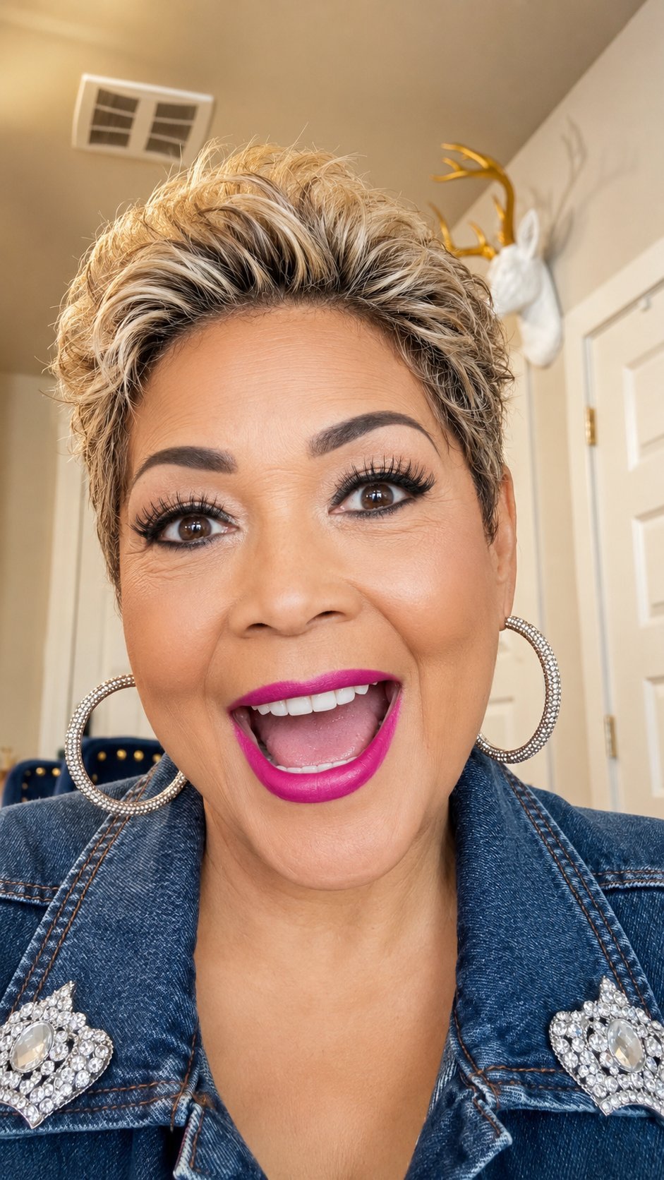 Woman with short blonde pixie hairstyle smiling at camera, wearing magenta lipstick, silver hoop earrings, and denim jacket with embellished shoulders