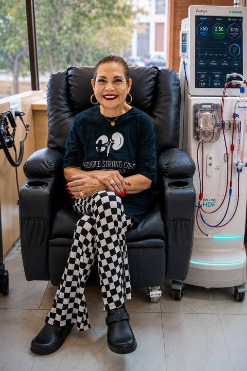 Woman smiling in dialysis treatment chair wearing kidney-themed shirt and checkered pants, with medical equipment beside her