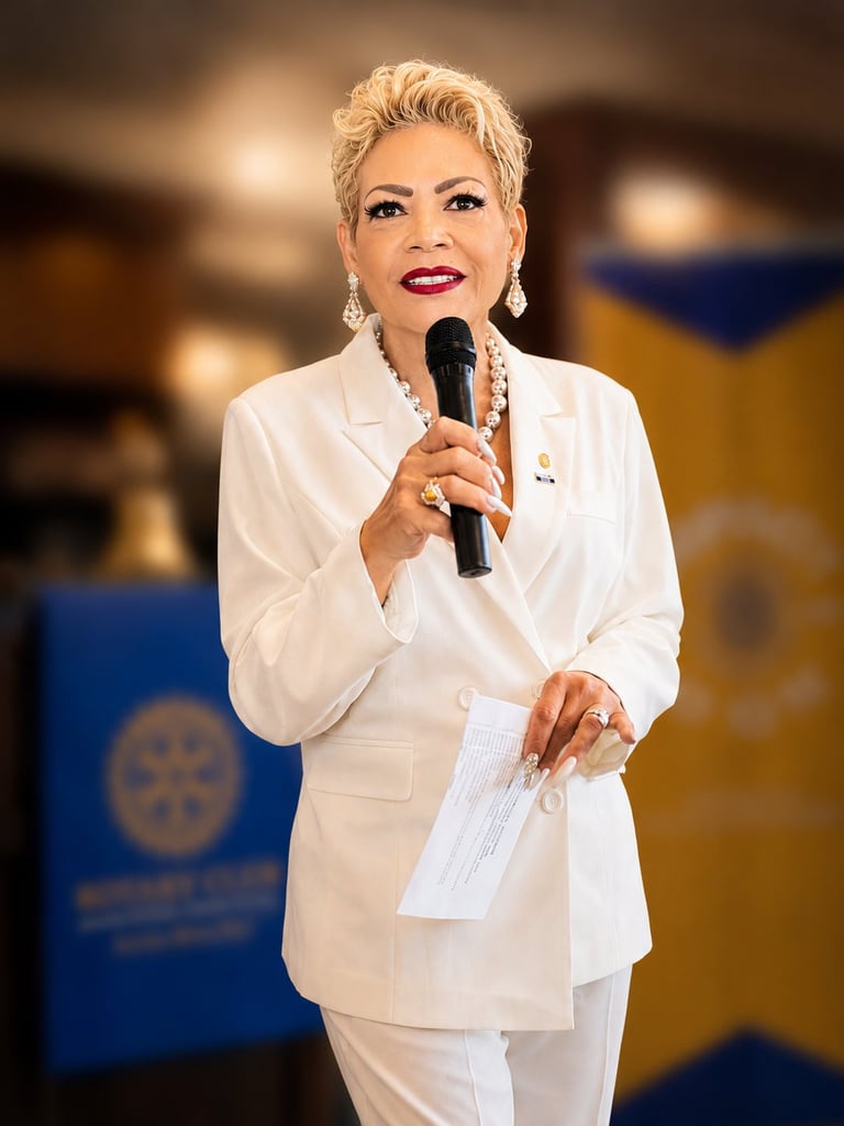 Woman with short blonde hair speaking at microphone wearing white blazer and pearl necklace