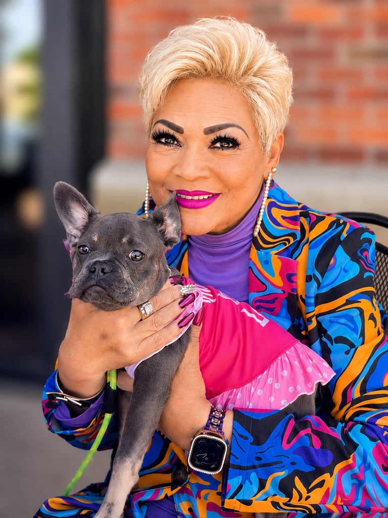Woman with short blonde hair and bright makeup holds a gray French Bulldog, wearing a colorful patterned jacket over a purple turtleneck