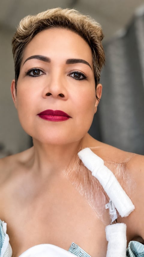 Headshot of a woman with short blonde hair, dark eyebrows, and magenta lipstick, wearing a white bandage on her shoulder