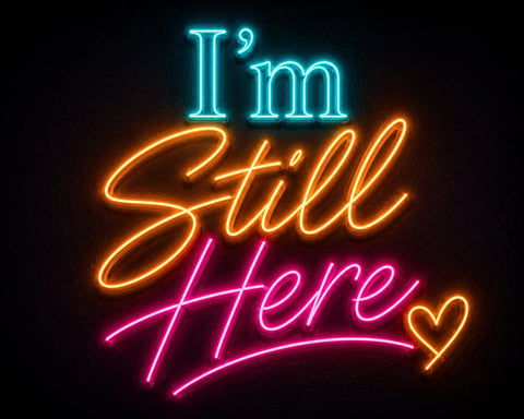 Neon sign reading Im Still Here in cyan, yellow, and pink glowing lights on a dark background