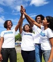 Group of volunteers in white shirts stacking hands together in unity outdoors with blue sky background