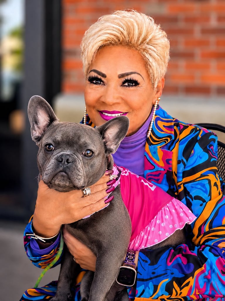 Woman with platinum blonde hair and vibrant makeup holding a gray French Bulldog, wearing colorful abstract print jacket and pink top