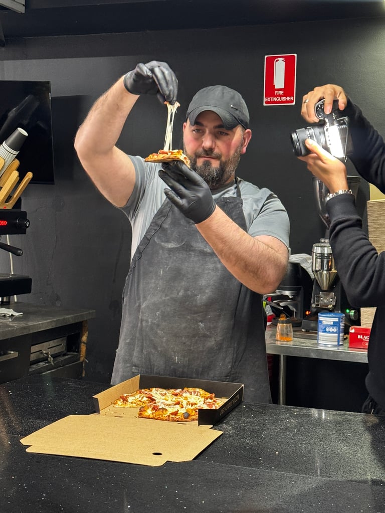 Man in gray apron and cap preparing pizza, stretching dough above a box with ingredients on counter