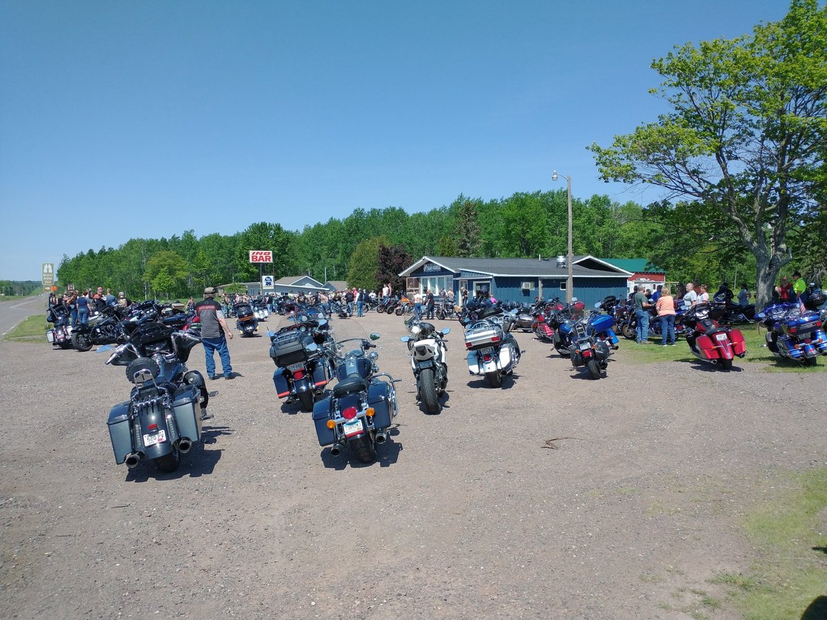 Large motorcycle rally gathering in a dirt lot with dozens of motorcycles parked and riders assembled near a building, surrounded by trees.