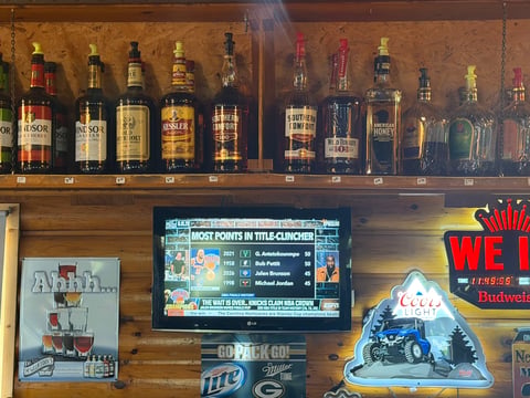 Wooden bar shelf displaying whiskey and spirit bottles above a sports scorecard display, neon signs, and beer branding