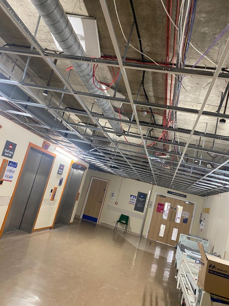 Industrial office interior with exposed ceiling ductwork, electrical conduits, and wiring above a concrete floor with elevator doors and storage boxes.