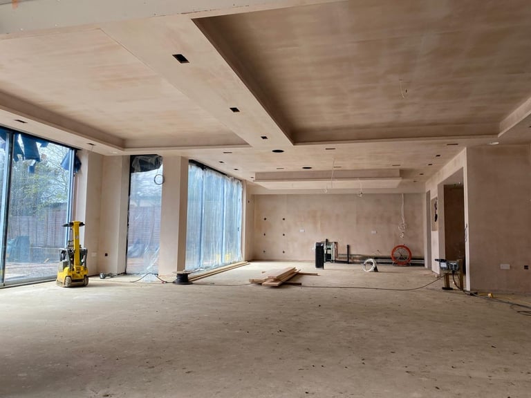 Empty construction site interior with drywall, recessed ceiling lights, and large windows overlooking outdoor area