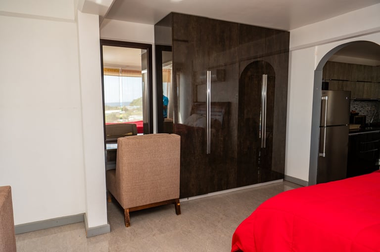 Modern bedroom with dark wood accent wall, upholstered chair, red bedding, and arched doorway leading to living area with windows