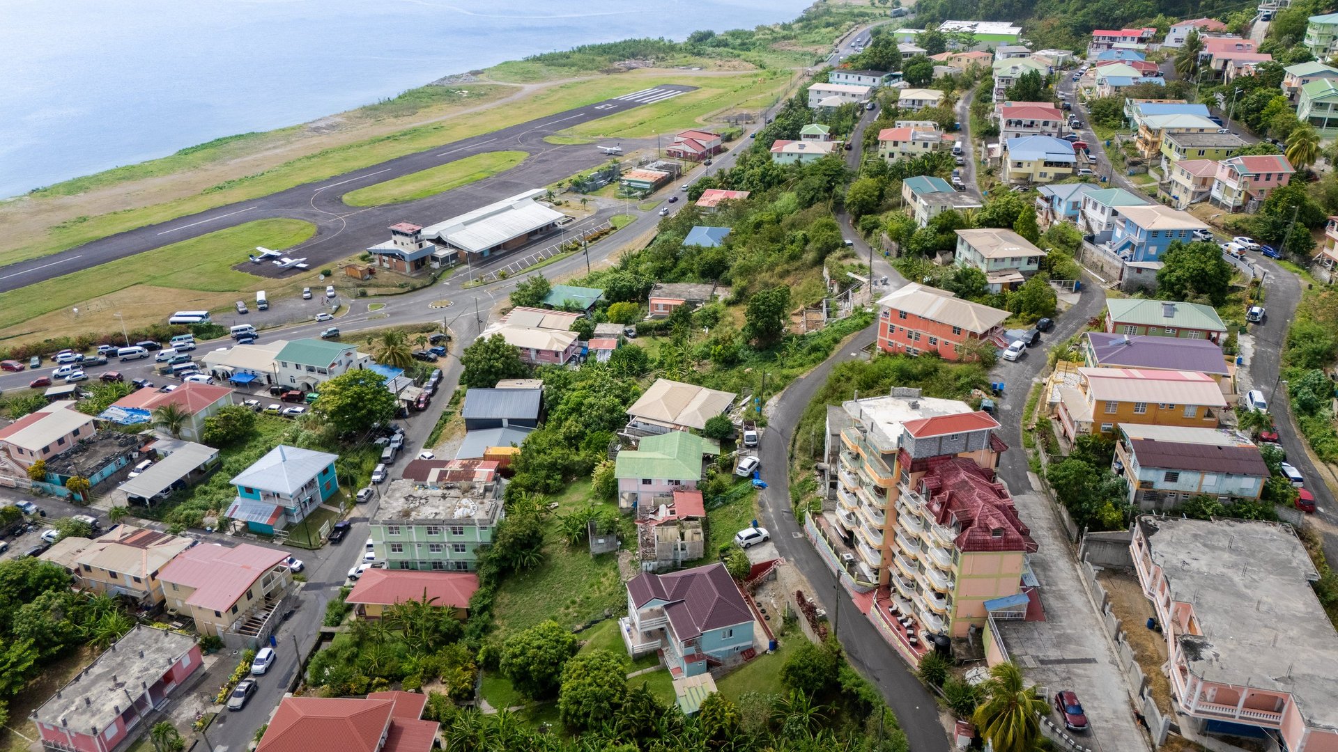 Aerial view of coastal town with airport runway, colorful houses, green trees, and water at Canefield, Dominica