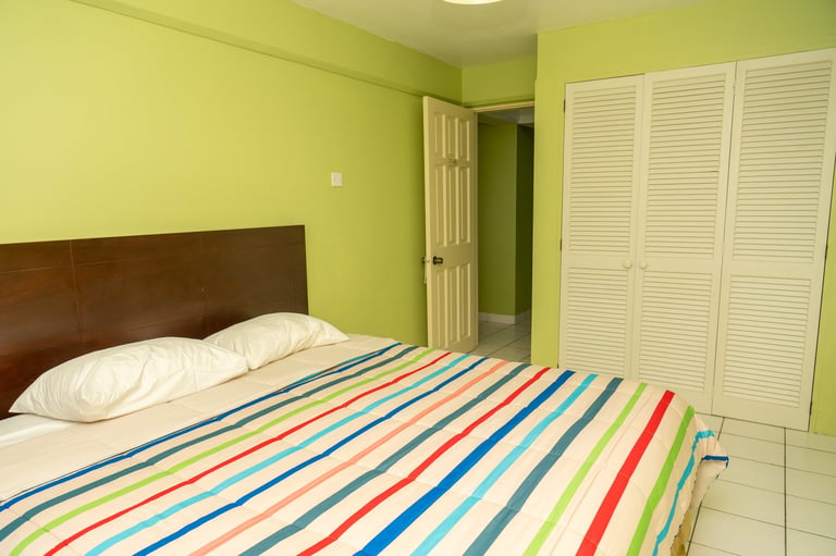 Bright lime green bedroom with brown headboard bed, colorful striped bedding, white louvered closet doors, and tile flooring