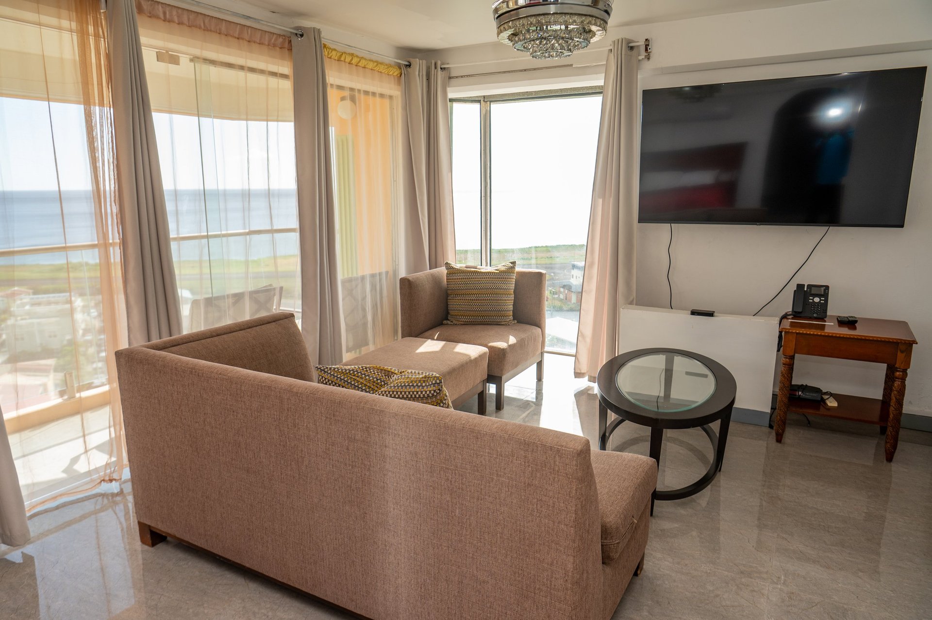 Modern high-rise Master Suite living room with beige sofa, ocean view windows, wall-mounted TV, and coastal furnishings overlooking the Caribbean Sea