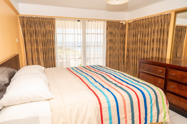 Spacious Triple Bedroom with colorful striped bedspread, white pillows, wooden furniture, and large windows with beige curtains