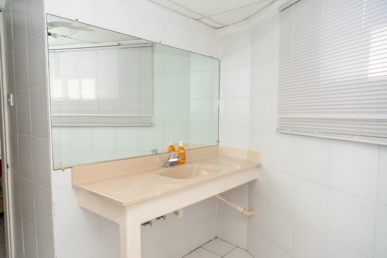 Minimalist bathroom workspace with beige desk, mirror, and white walls lit by natural light from window blinds