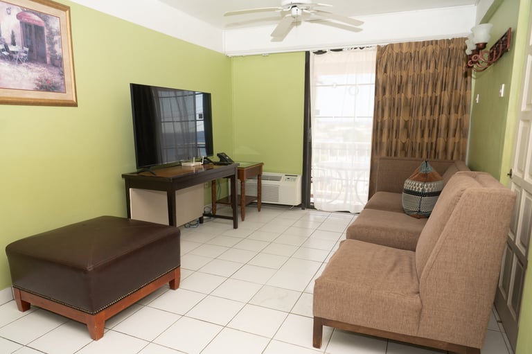 Bright yellow living room with brown sofa, ottoman, flat-screen TV on stand, tiled floor, and glass doors to patio