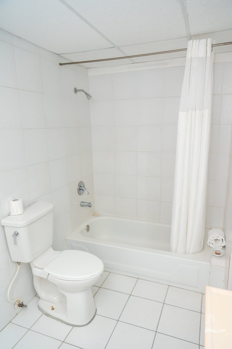 A clean, minimalist bathroom with white tile walls, a toilet on the left, and a bathtub with white shower curtain on the right
