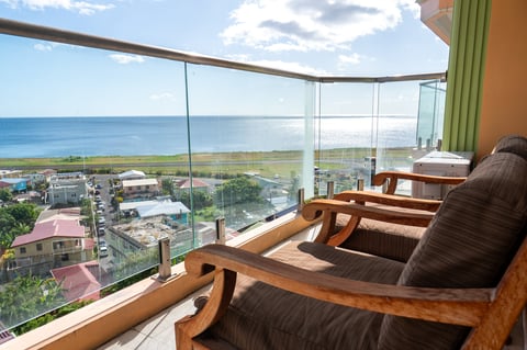 Balcony seating area with panoramic ocean view through floor-to-ceiling windows overlooking tropical hillside and sea